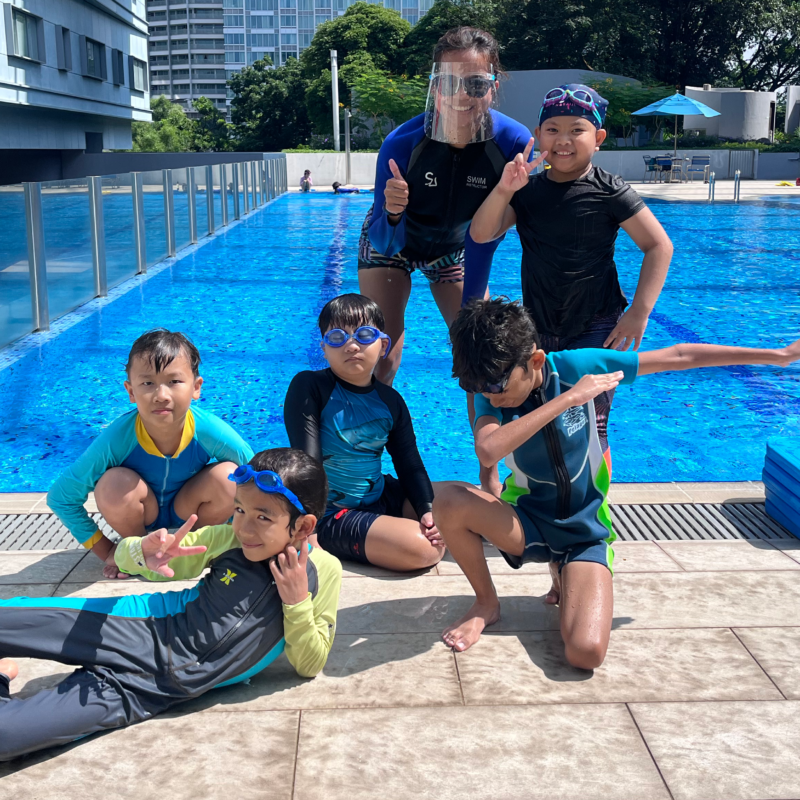 Group of children smiling in front of Swim Lab pool, representing different age groups and swimming levels.