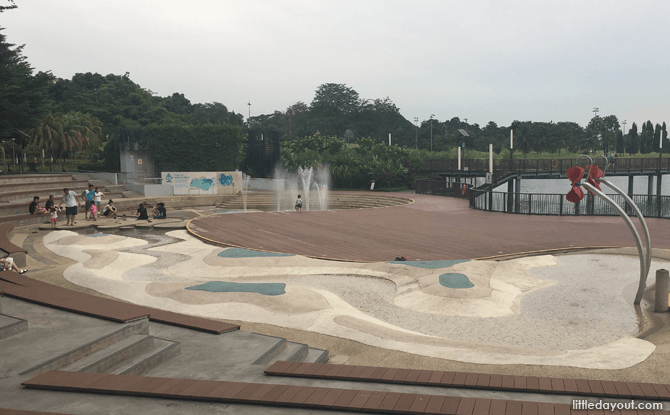Open-air playground near a reservoir with water sprouts scattered around, offering space for children to run and parents to relax.